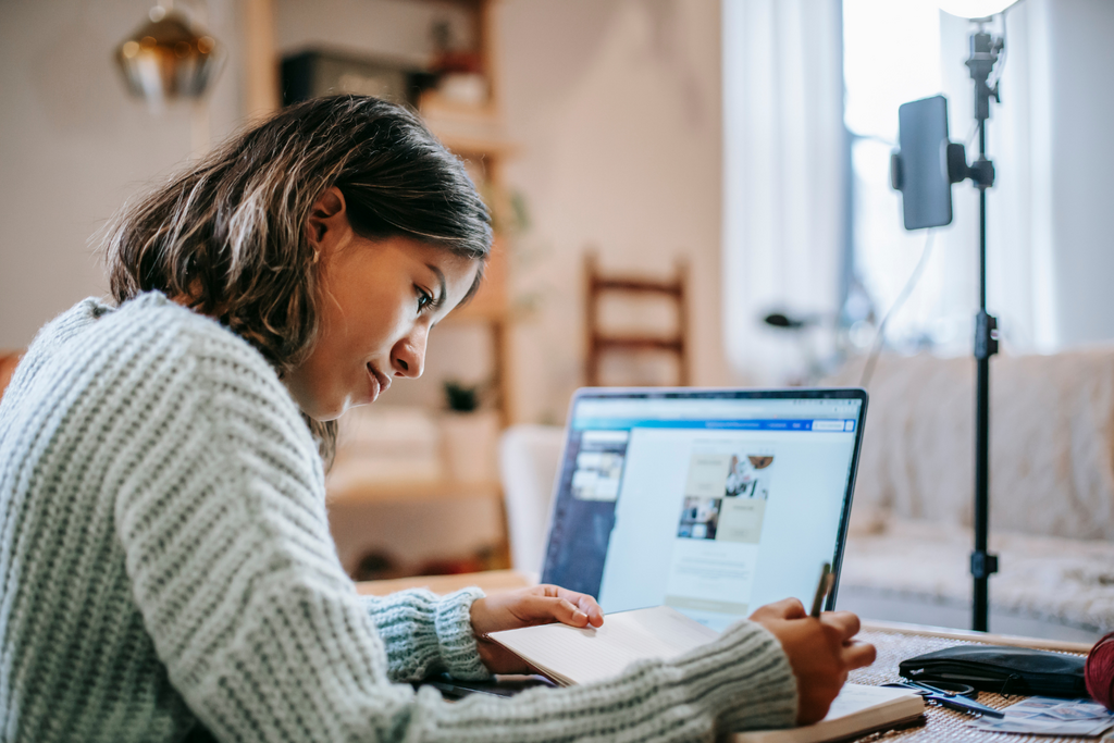 Student sat at desk in her room on laptop and writing in notebook